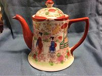 Side view showing hand painted Asian figures and floral designs on porcelain tea pot with red handle and spout.