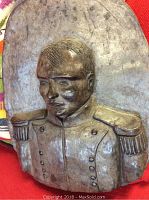 Front view of bronze or metal relief sculpture bust of Napoleon Bonaparte on red fabric background showing head and shoulders.