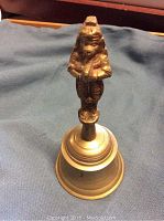 Top-down photo of the brass bell placed on a blue fabric, showing the figure on the handle and the bell base with number '2'.