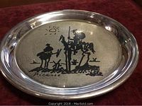 Front view of round sterling silver plate with black line drawing of Don Quixote and Sancho Panza under sun, signature “Picasso”