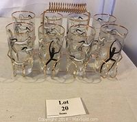 Front view of all eight glass tumblers held in the gold-toned metal carrying rack showing black dancing figure designs and gold trim.