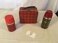 Metal lunchbox with matching red plaid thermos and an additional green patterned thermos, all on a white fabric background.