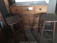 Photo showing natural wood nine drawer dresser with round wooden knobs and two tall wooden barstools with square seats beside it. Wear and staining visible on all items.