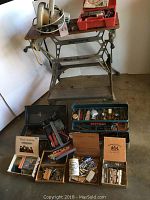 Full view of Black and Decker Workmate adjustable workbench with circular saw on top and assortment of toolboxes and cigar boxes of hardware on the floor in front.