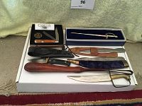 Full lot of knives displayed on white box showing decorative dagger, Camillus pocket knife in box, several fixed blade knives with sheaths and throwing knives.
