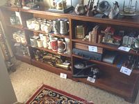 Two wooden bookshelves filled with various items displayed side by side, with a corner shelf next to them. Shelves contain mugs, small decorative objects, books, and other items, showing the scale and wear of the shelves.