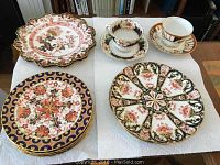 Overview of eight plates and two teacup-and-saucer sets displayed on white protective padding in front of bookshelf.