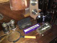 Tabletop with brass opera glasses, eyeglasses in case, magnifying glass, purple box with fountain pen, clear plastic case with two marbled pens, second pair of binoculars partially visible, wooden barometer/clock plaque above