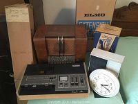Overview of assorted vintage radios, movie reel box, screen, and clock showing the lot's contents