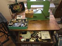 Green Elna sewing machine on wooden table with metal extension and open drawer of spools and accessories.