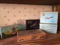 Seven assorted vintage cigarette tins and cases displayed on wooden shelf, showing varied designs and wear.