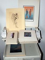 Overall view of collection showing multiple matted prints stacked and displayed on white furniture, including a black and white winged figure sketch and color prints of cloaked figures.