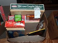 Photo showing multiple board games and craft kits in a cardboard box with visible labels for Flower Candle Maker candle kit, Resin Art kit, Mini Master Mind, alphabet cards, and Joker games.