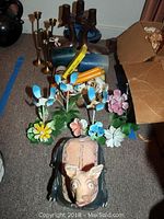 The full lot showing brass candlesticks, ceramic pig planter, colorful metal flowers, and blue decorative glass dish spread on the floor next to a cardboard box.