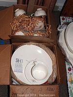 Photo of dinnerware plates, bowls, and cups packed in a box with shredded packing paper, showing they are new and not used.