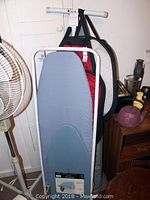 Photo of three ironing boards with covers in blue, red, and black-grey colors, leaning against wall with laundry bags attached by straps.