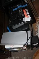 Stacked electronics including a black GE VHS VCR, a black boombox, and a black CRT box television underneath a silver Toshiba DVD player, all on a carpeted floor.