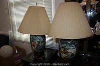 Two Asian style ginger jar ceramic table lamps with beige pleated fabric shades on a glass surface in front of a window. The shades show wear and staining.