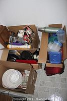 Overview of boxes containing assorted used kitchenware including frying pans, kitchen utensils, plastic containers, and plates.