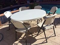 Oval white heavy duty plastic folding table surrounded by six matching white folding chairs with metal frames on tiled patio by pool.