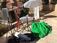 Array of photography lighting equipment including Husky dual-head work lights on a tripod, two white umbrellas on stands for light diffusion, green fabric backdrop or green screen, and light bulbs on the ground outdoors.