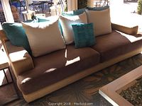 Outdoor sofa with tan frame and brown cushions, accompanied by several beige and teal pillows. Fabric shows slight fading.