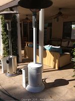 Full view of the white outdoor heater showing cylindrical body, base door open, and tall chimney with reflector top in patio setting.