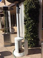 Outdoor patio heater in tall white metal housing with circular base and vented panels, showing wear and rust spots, standing on outdoor tile surface near building and greenery.