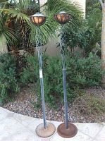 Two tall outdoor oil torches standing side by side among garden plants with twisted wrought iron poles and copper-colored oil reservoirs at the top.