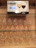 Shelf showing 19 used clear wine glasses of various sizes arranged in rows and one box of Symphony Break-Resistant wine glasses on the top shelf.