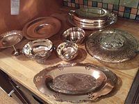 Full view of all eight silver plated serving pieces displayed on wooden surface, showing trays, bowls, and circular pieces with ornate designs.
