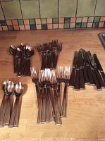Full set of Lenox stainless steel cutlery items arranged on wooden surface, showing all forks, spoons, and knives organized in groups.