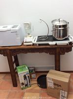 View of all lot items arranged on a wooden table and floor: Breadman bread machine, Osterizer blender, Toastmaster griddle, Agostina pot, boxed Ninja blender, boxed Sodastream machine, and black frying pan.