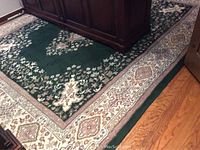 Top view of a large green area rug with floral design and beige border in a room with wooden floor and furniture.