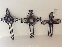 Three wrought iron crosses hanging on a white wall, showing overall view, sizes, and decorative details.
