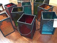 Eight outdoor lanterns displayed on a wooden surface, showing various colors and black metal frames with handles.