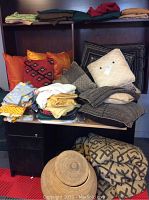 Full view of bed linens, pillows, table linens stacked on a black desk with red and black checkered floor tiles.