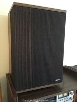 Front view of one speaker showing black fabric grille and wood-grain vinyl cabinet.