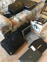 View of all electronics including printer, scanner, laptops, laptop case, and box of cables on couch and carpet.