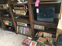 Three wooden shelf units with four shelves each, filled with audio equipment, vinyl records, and other media items. Roll-up wooden doors meet in the middle of each unit.