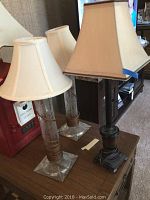 Three table lamps on a wooden surface. Two lamps have crystal bases and cylindrical clear bodies with white fabric shades. The third lamp has a dark wooden body with a square beige fabric shade.