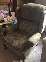 Wingback recliner chair with patterned fabric upholstery and tack accents, showing wear on cushions.