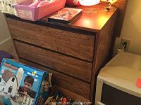 Wooden dresser shown with three drawers and visible wood grain, alongside some household items.