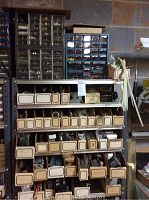 Display of labeled cardboard boxes on metal shelves containing various parts including screws, transistors, and grommets.