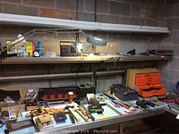 Full view of a workbench in a basement with a variety of hand tools, hardware, and electronic components arranged on its surface and shelves.