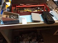 Full view of several tools in a red metal organizer tray atop a table, next to two small boxes with drill bits and hardware items