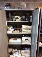 Metal storage cabinet containing multiple computer parts, manuals, and items stored in labeled boxes on shelves.