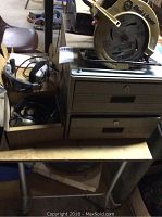 Gray metal 2 drawer file cabinet with box and cables on top on metal folding table.