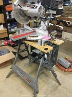 View of the miter saw mounted on a foldable Black & Decker work table. The saw blade and table legs are visible.