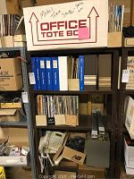 Photo of shelving unit with blue binders, magazines, a box labeled 'OFFICE TOTE BOX' containing model train parts, a rotary phone, and metal items on lower shelf.